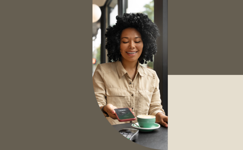 Mulher negra sorri enquanto utiliza o celular para realizar um pagamento em uma maquininha. À sua frente, na mesa, há uma xícara de café azul.